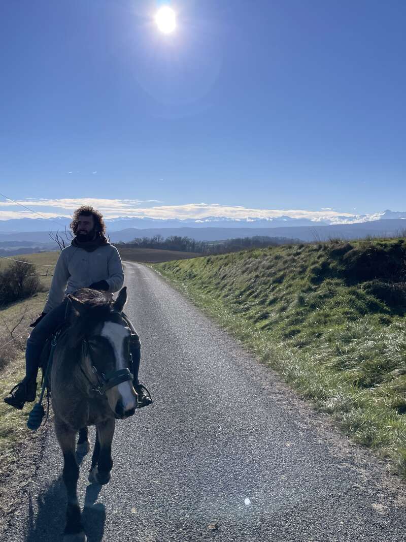 Un hombre de pelo rizado cabalga por un camino rural en un día soleado. Las majestuosas montañas y el cielo azul crean un impresionante telón de fondo.