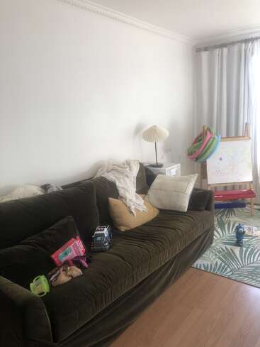 A cozy living room corner with a dark green velvet sofa, scattered toys, children’s books, pillows, a lamp, easel, colorful scarves, and leafy patterned rug.