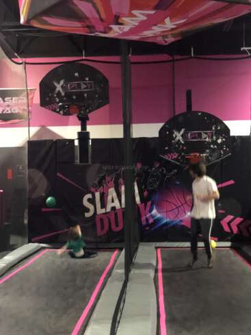 The image shows two people in an indoor trampoline park, one jumping and another falling. Basketball hoops and colorful "Slam Dunk" signage are visible in the background.