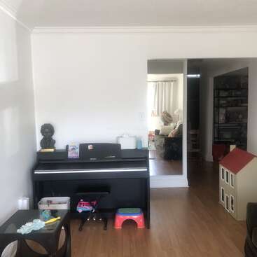 This image shows a cozy living room with a black piano, a bust sculpture, children's toys, a dollhouse, wooden floors, and a peek into an adjoining room.