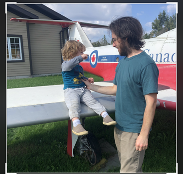 Un homme et un enfant discutent à côté d'un avion des Forces canadiennes. L'enfant est assis sur l'aile, les deux sourient, avec un bâtiment et de l'herbe verte à proximité.