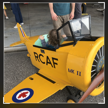 Un jeune enfant est assis à l'intérieur d'une petite maquette d'avion jaune portant l'inscription "RCAF MK II", tandis que des adultes se tiennent à proximité. Le décor semble être un hangar intérieur.