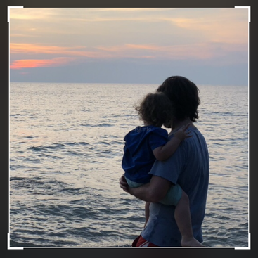 Une personne tient un enfant dans ses bras au bord de la mer au coucher du soleil, créant un moment de sérénité et d'affection sur fond d'eau calme et de ciel coloré.