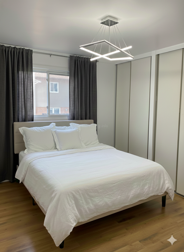 Une chambre à coucher minimaliste comprend un lit bien fait avec des draps blancs, un plafonnier géométrique moderne, de grandes armoires, un plancher en bois et des rideaux gris couvrant une fenêtre.