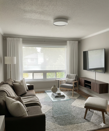 Un salon moderne aux tons neutres comprend un canapé marron, une table basse en verre, un fauteuil, une grande fenêtre avec des rideaux blancs, une télévision murale et une décoration élégante.