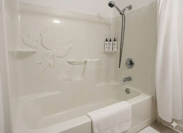 Cette image montre une baignoire blanche et propre avec un motif floral sur le mur, une pomme de douche, trois bouteilles, une serviette et un rideau de douche blanc.