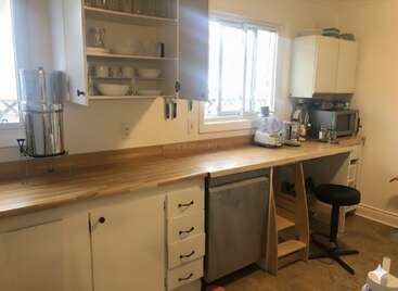 Cette cuisine comprend des comptoirs en bois, des armoires blanches, des étagères ouvertes avec de la vaisselle, un filtre à eau, un mixeur, un micro-ondes, une fenêtre et un tabouret sous un espace de travail.