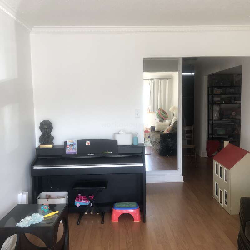This image shows a cozy living room with a black piano, a bust sculpture, children's toys, a dollhouse, wooden floors, and a peek into an adjoining room.