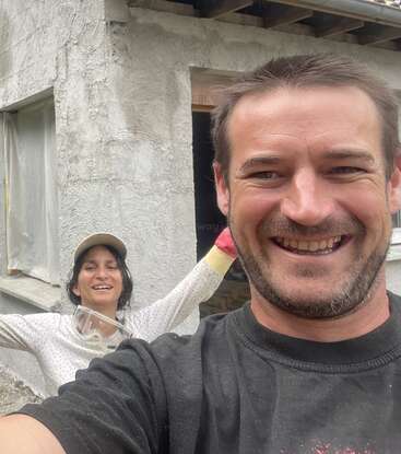 Zwei Menschen lächeln fröhlich vor einem Haus, das gerade renoviert wird. Eine Person posiert mit ausgestreckten Armen, während die andere ein fröhliches Selfie aus der Nähe macht.
