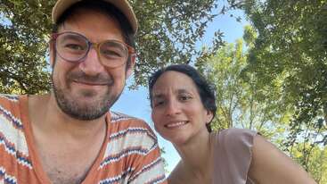 Ein Mann und eine Frau lächeln draußen unter laubreichen Bäumen in die Kamera. Der Mann trägt eine Brille, eine Mütze und ein gestreiftes Hemd; die beiden sehen glücklich aus.