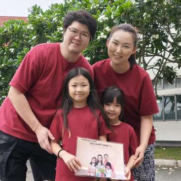 Une famille de quatre personnes, toutes vêtues de chemises rouges assorties, pose à l'extérieur en souriant. Les deux jeunes filles tiennent devant elles une photo de famille encadrée.