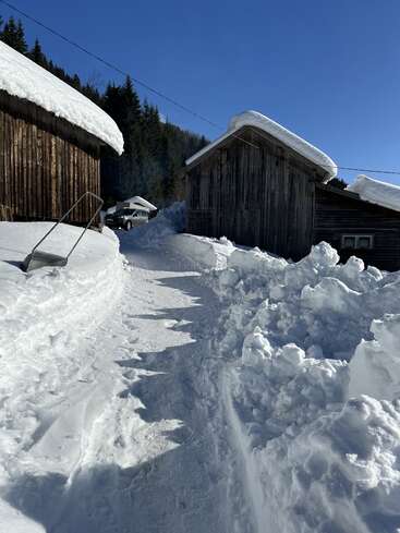 Ein schmaler Pfad schlängelt sich zwischen hölzernen Hütten, die mit dichtem Schnee bedeckt sind. Der Himmel ist klar und blau, im Hintergrund stehen hohe Kiefern und ein Auto ist teilweise zu sehen.