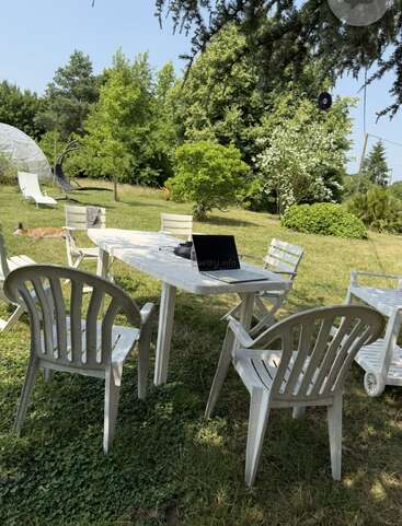 Plastic chairs surround a white outdoor table with a laptop on it. The setting is a sunny, green garden with trees, bushes, and a relaxed vibe.