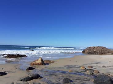 Uma cena de praia deslumbrante com areia dourada, pedras espalhadas, ondas suaves batendo e um céu azul claro. A atmosfera tranquila é pacífica e convidativa.