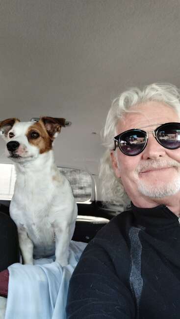 A cheerful man with white hair and sunglasses takes a selfie in a car, accompanied by a cute, alert brown and white dog sitting beside him.