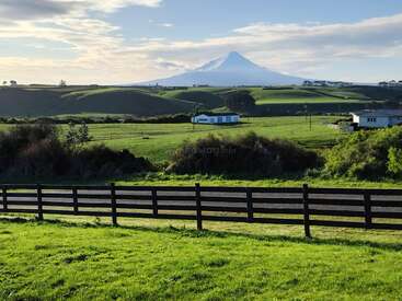 A lush green field with black fencing, a few houses, rolling hills, and a majestic snow-capped mountain in the background under a partly cloudy sky.