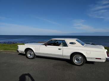 A classic white car is parked near a scenic coastal road, with calm ocean waves and a clear blue sky in the background on a sunny day.