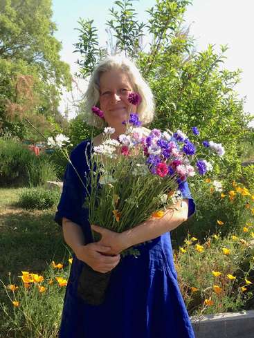 L'image représente une femme âgée debout dans un jardin, tenant un vibrant bouquet de fleurs sauvages, vêtue d'une robe bleue, dans une atmosphère sereine et paisible.