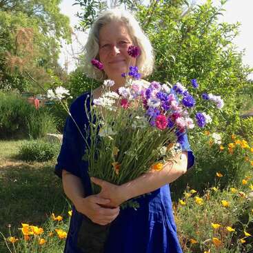 Das Bild zeigt eine ältere Frau, die in einem Garten steht, einen bunten Strauß Wildblumen in der Hand hält und ein blaues Kleid trägt, mit einer ruhigen und friedlichen Atmosphäre.