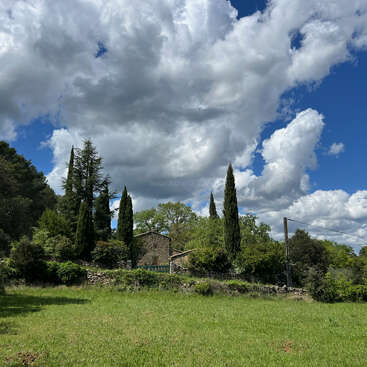 A rustic stone house is nestled among tall cypress trees and lush greenery, under a dramatic sky filled with large, white cumulus clouds and vibrant blue patches.