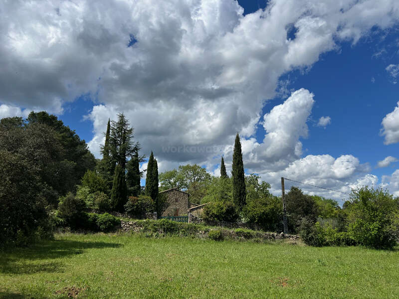 Entre altos cipreses y exuberante vegetación se alza una rústica casa de piedra, bajo un espectacular cielo repleto de grandes cúmulos blancos y vibrantes manchas azules.