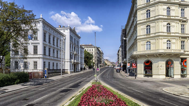 L'image représente une rue de ville avec une rangée de bâtiments, une route avec un trottoir et une zone herbeuse, avec quelques arbres et un plan d'eau à l'arrière-plan.