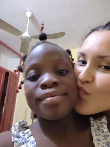 Dieses Bild zeigt zwei Mädchen, die eng beieinander stehen und sich gegenseitig einen Kuss auf die Wange geben. Sie sehen glücklich aus, und im Hintergrund ist ein Deckenventilator zu sehen.