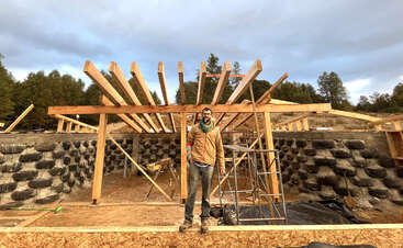 The image shows a construction site with a building under development, featuring a wooden frame and a concrete foundation, surrounded by a grassy area with trees and a clear sky.