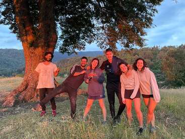 L'image représente six personnes debout dans un champ herbeux, avec un grand arbre derrière elles et un paysage montagneux à l'arrière-plan sous un ciel bleu.