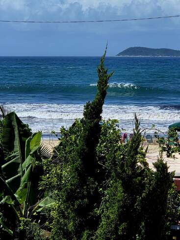 Árboles y plantas de un verde exuberante enmarcan la vista de una playa de arena y las olas de un océano de un azul intenso, con una isla en la lejanía bajo un cielo parcialmente nublado.