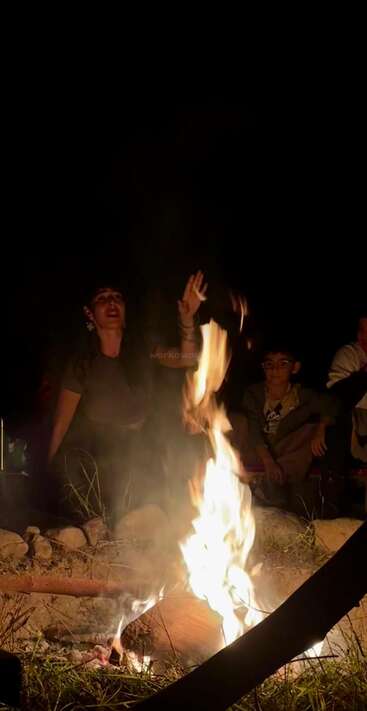 Un groupe de personnes est assis autour d'un feu de camp la nuit. Une personne fait des gestes animés tandis que les autres écoutent, créant ainsi une atmosphère chaleureuse, communautaire et détendue en plein air.