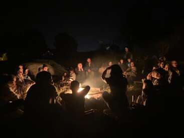 Un groupe de personnes est assis la nuit autour d'un feu de camp rougeoyant, profitant de la compagnie, de la conversation et de la chaleur des autres sous un ciel sombre et étoilé.