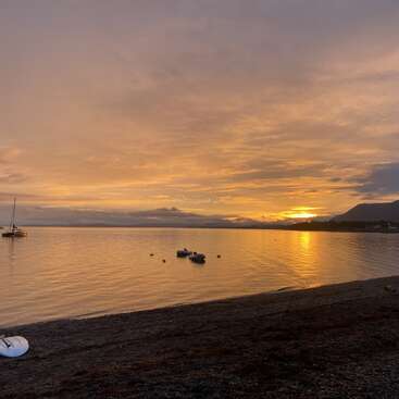 The image depicts a serene beach scene at sunset, featuring a calm body of water, a sandy shore, and a few boats, with a mountain range visible in the distance.