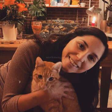 The image depicts a woman with long dark hair, cradling an orange tabby cat, set against a cozy background of a brick wall and wooden table.