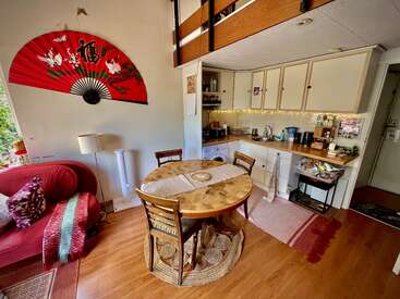 The image depicts a cozy living room and kitchen area, featuring a wooden dining table, red couch, and white cabinets, with a large fan on the wall and a doorway leading to the outside.