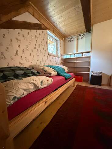 Dieses gemütliche Dachgeschoss-Schlafzimmer hat ein Einzelbett mit bunter Bettwäsche, verspielte Tiertapeten, Holzwände, Regale, Kleiderbügel, ein kleines Fenster und einen tiefroten Teppich.