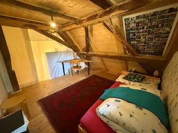 Dieses gemütliche Schlafzimmer im Dachgeschoss hat freiliegende Holzbalken, eine schräge Holzdecke, ein Einzelbett mit gemusterter Bettwäsche, einen roten Teppich, einen kleinen Schreibtisch, einen Stuhl und eine Fotocollage.