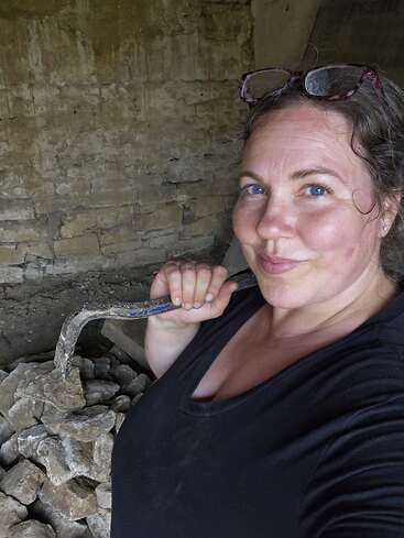Uma mulher com óculos na cabeça, segurando um pé de cabra, está em frente a uma pilha de pedras contra uma parede de pedra áspera e inacabada, sorrindo com confiança.