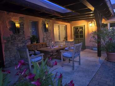 Pátio externo aconchegante com móveis rústicos de madeira, mesa de jantar à luz de velas, vasos de plantas, paredes de pedra e laranja, iluminação suave à noite e ambiente convidativo sob um telhado coberto.