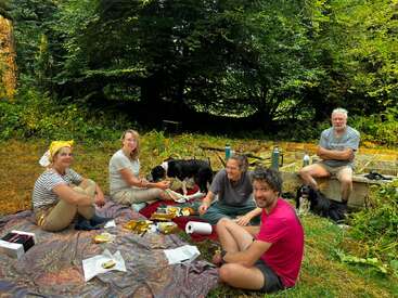 Fünf Menschen genießen ein Picknick im Freien in einer üppigen, grünen Umgebung. Zwei Hunde begleiten sie. Essen, Getränke und ein entspanntes Lächeln deuten auf ein fröhliches, friedliches Beisammensein hin.