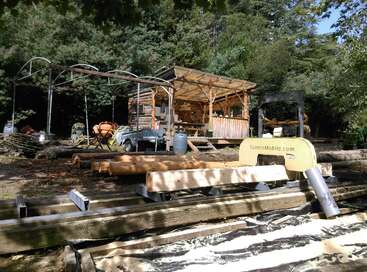 La imagen representa un rústico taller al aire libre, con una máquina amarilla, vigas de madera y una estructura cubierta, rodeada de árboles y un sereno entorno natural.