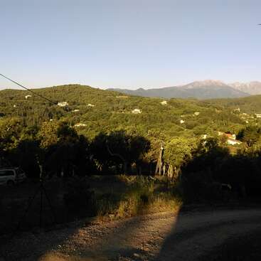 La imagen muestra un paisaje sereno con un camino de tierra, árboles frondosos y montañas lejanas, con un cielo azul despejado y algunas casas enclavadas entre la vegetación.