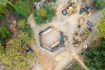 La imagen representa una obra en construcción con una gran zona abierta en la que hay un montón de madera y un edificio parcialmente construido, rodeado por una valla y un camino de tierra.