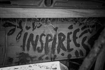 La imagen muestra un muro con la palabra "inspire" en letras blancas, acompañada de una pequeña zona verde y una base de hormigón gris. El muro está situado frente a una masa de agua.