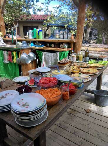 La imagen muestra una cocina rústica al aire libre con una mesa de madera cargada de una gran variedad de platos, cuencos y alimentos, sobre un sereno fondo natural.