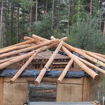 La imagen representa un edificio en construcción, con un tejado de troncos de madera y una pared de barro o arcilla, con árboles visibles al fondo.