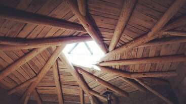 La imagen representa una estructura de madera con tejado inclinado, con una viga prominente y una serie de vigas más pequeñas, probablemente una casa o un edificio, con un entorno natural y un cielo despejado.