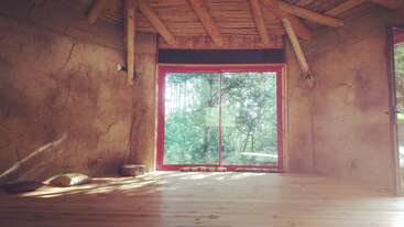 La imagen muestra una habitación con suelo y paredes de madera, una mesa y sillas, una ventana a la izquierda y una puerta a la derecha, todo ello en un tono neutro.