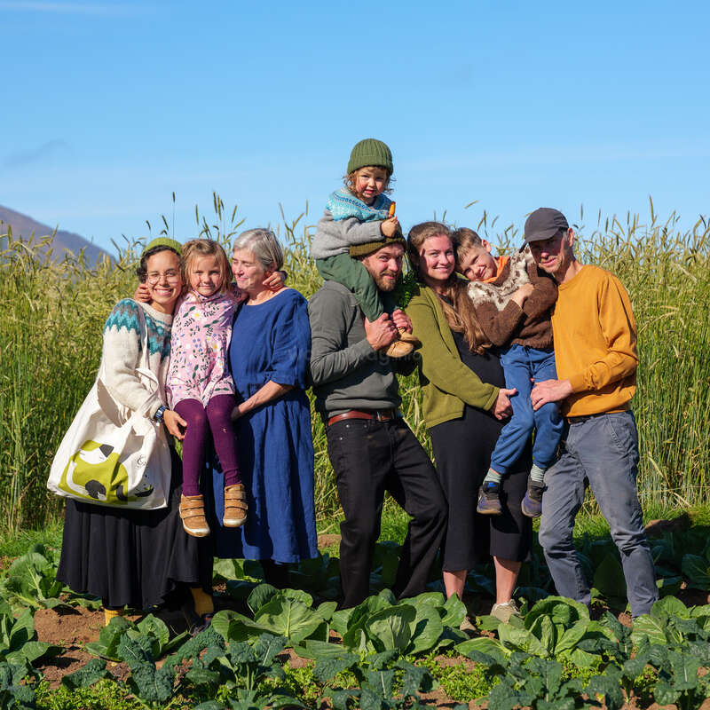 Une joyeuse famille de huit personnes, adultes et enfants, pose ensemble dans un potager luxuriant sous un ciel bleu clair, profitant d'une journée ensoleillée dans la nature.