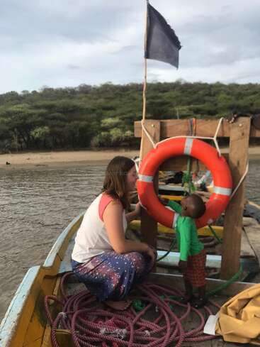 A woman and a child are on a small boat in the water, with a life ring and rope visible, set against a scenic background of trees and a cloudy sky.